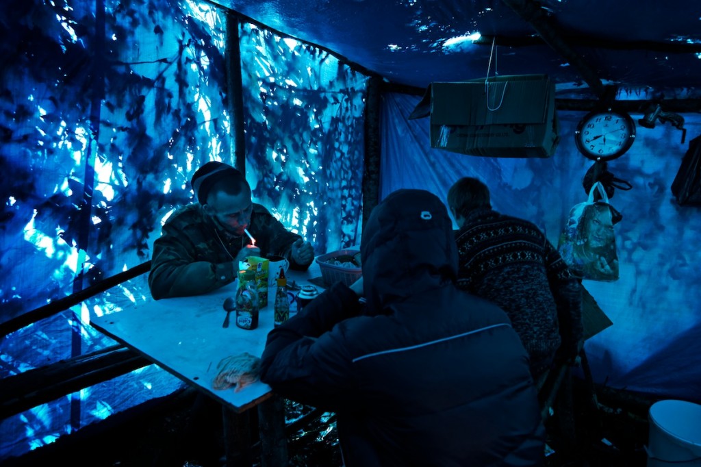 Blue light inside tent with man lighting cigarette at a table with two other men