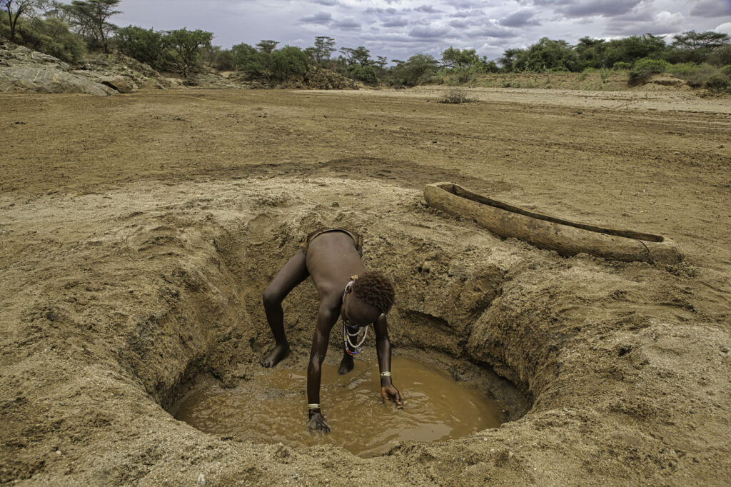 child reaches into muddy water in a small pit in barren landscape